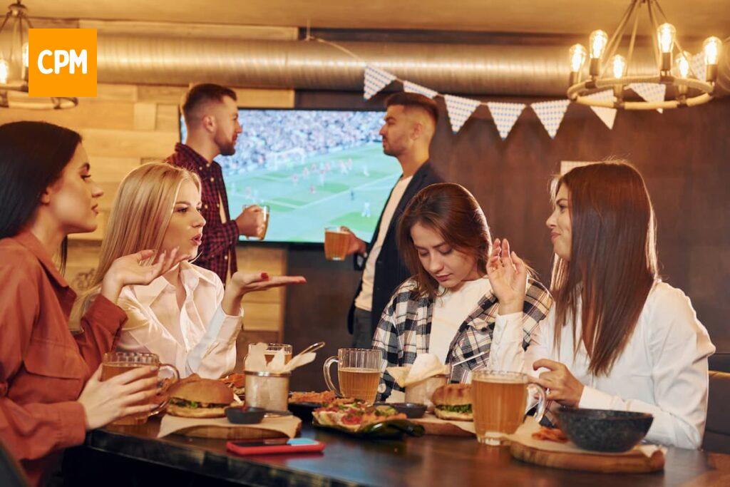 Imagem mostra grupo de amigos assistindo jogo, bebendo cerveja e lanchando.
